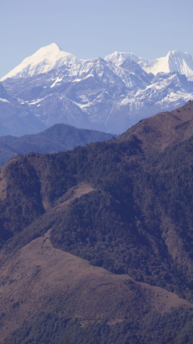 Snow-capped mountains rise majestically in the background, with layered green hills and valleys in the foreground, showcasing a stunning natural landscape.