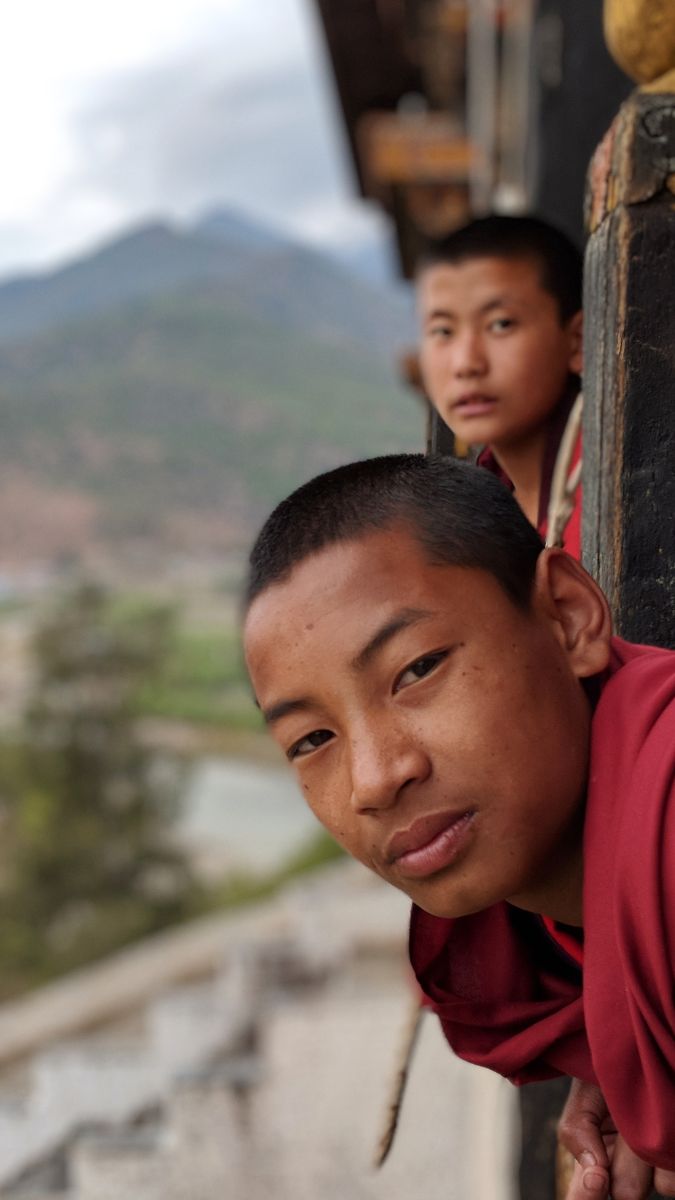 Two young monks in crimson robes peer out from a window. The background features a scenic landscape with mountains and greenery. One monk is closer to the camera, with a serious expression, while the other is slightly behind, looking at the viewer.