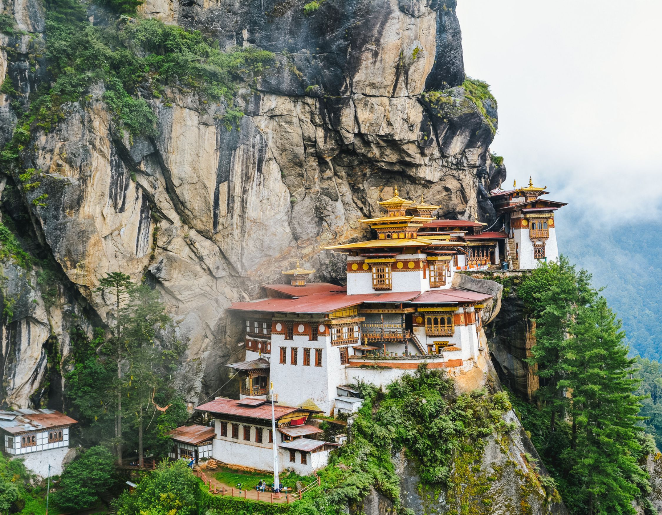 A striking mountain cliffside monastery with traditional architecture nestled amongst lush green trees and fog. The structure features ornate roofs, wooden balconies, and is surrounded by dramatic rock formations.