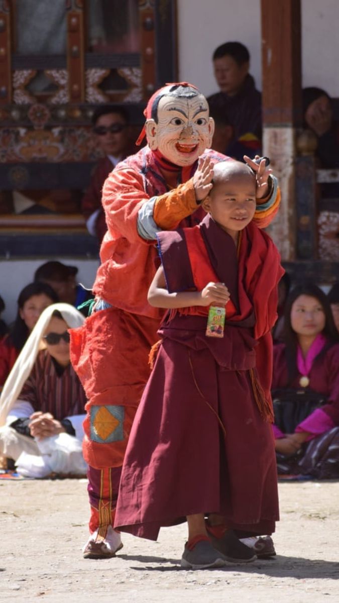 A traditional masked performer in colorful attire interacts playfully with a young monk in red robes during a cultural festival, with an audience watching in the background.