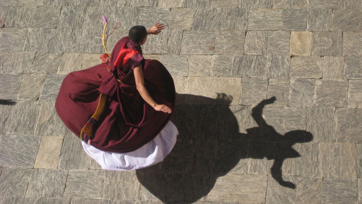A dancer in traditional attire spins gracefully on a stone pavement, casting a long shadow. The vibrant colors of the clothing contrast with the grey stones below.