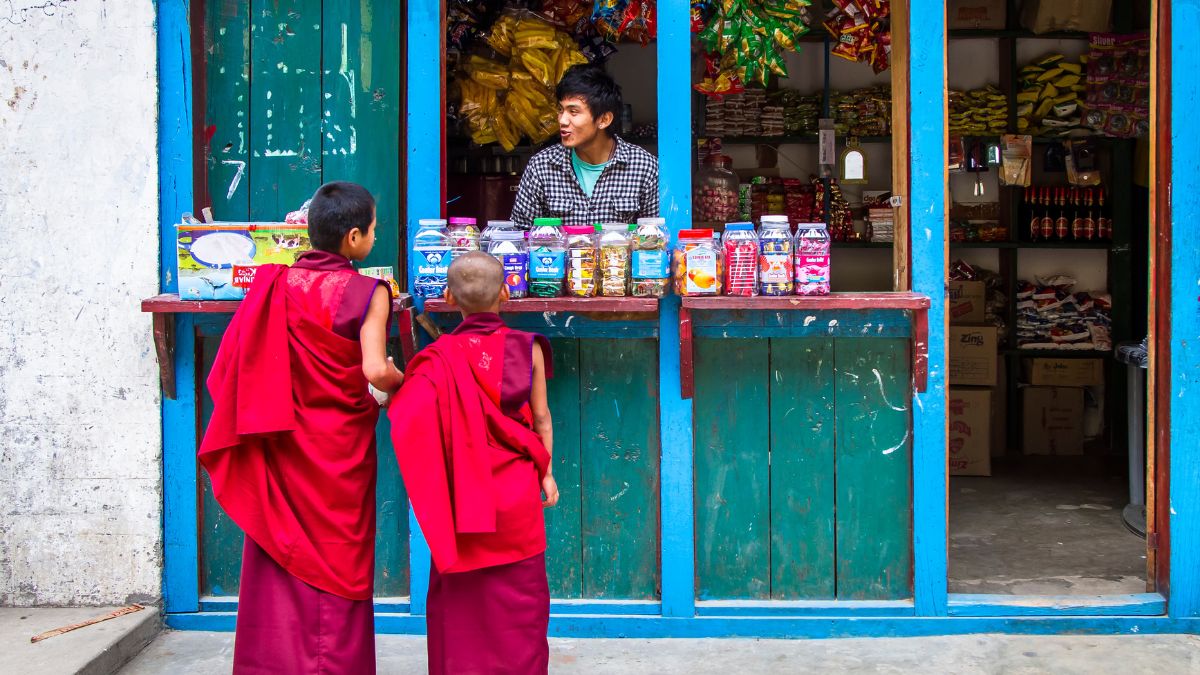 Two young boys in red monk robes stand in front of a small shop, engaging with a young man behind the counter. The shop displays a variety of goods in glass jars and colorful packaging.