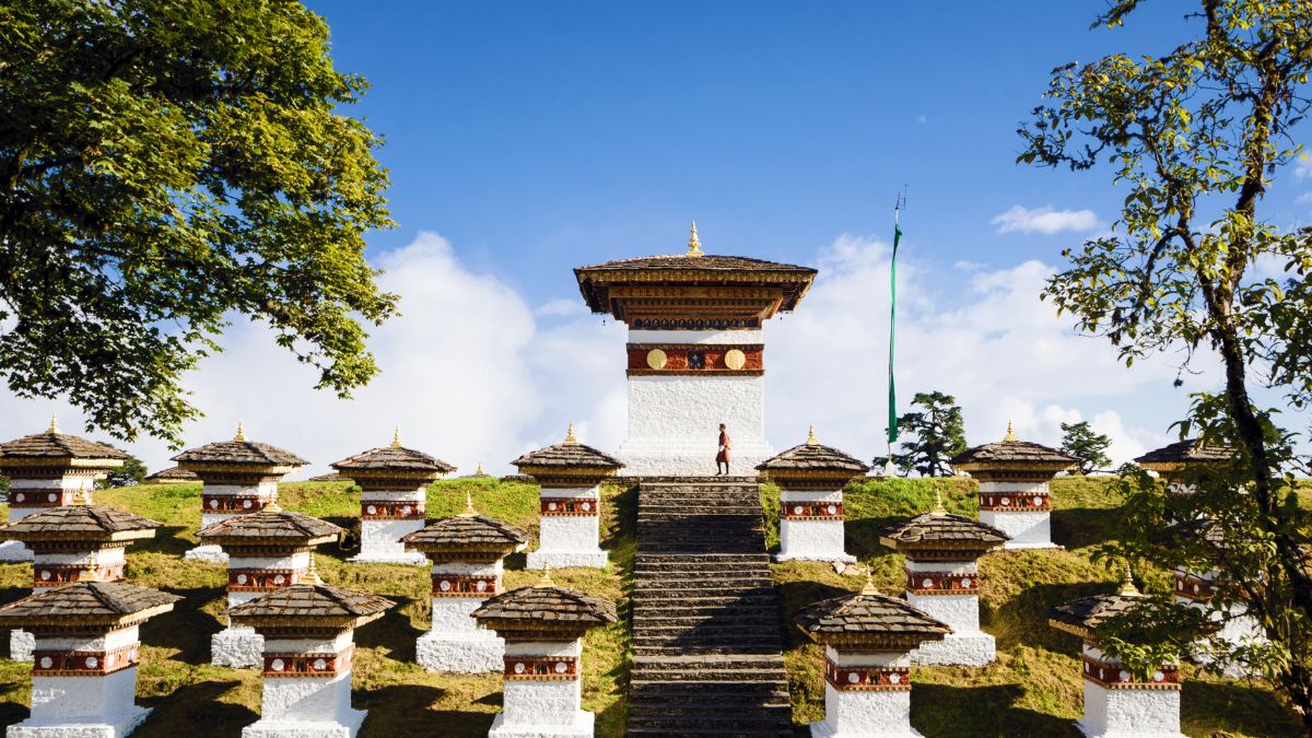 A serene landscape featuring a series of white stupas with ornate roofs, set against a clear blue sky. A person stands on the steps of the central stupa, surrounded by lush greenery.
