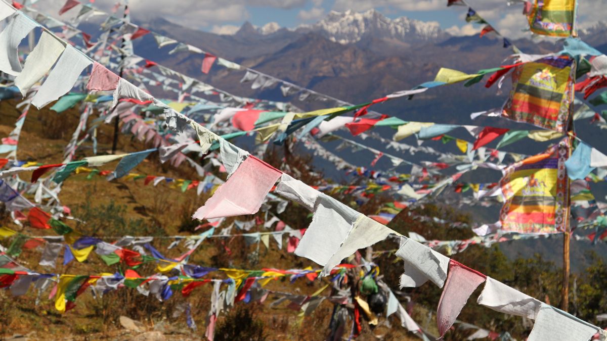 Colourful prayer flags flutter in the wind against a backdrop of snow-capped mountains and a clear blue sky, creating a vibrant and serene atmosphere.