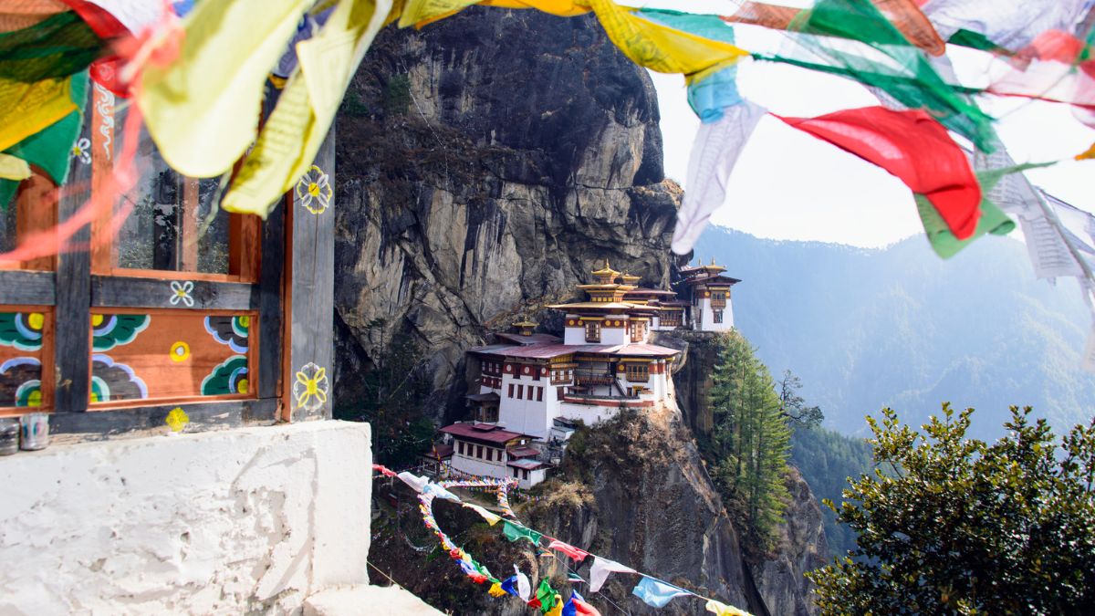 A breathtaking view of the Paro Taktsang monastery, perched on a cliffside in Bhutan, surrounded by lush mountains. Colorful prayer flags flutter in the foreground.