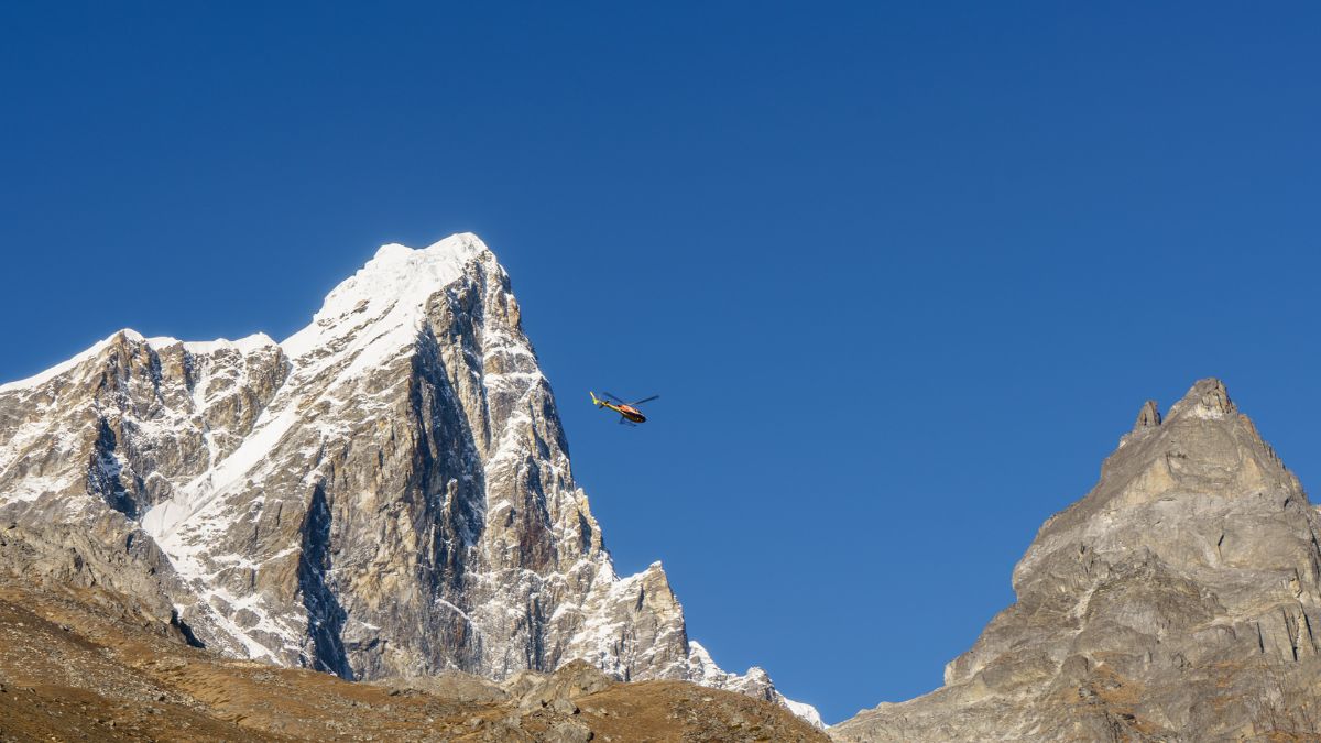 A helicopter flies above a snow-capped mountain against a clear blue sky. Another rugged peak is visible in the foreground, showcasing a dramatic alpine landscape.