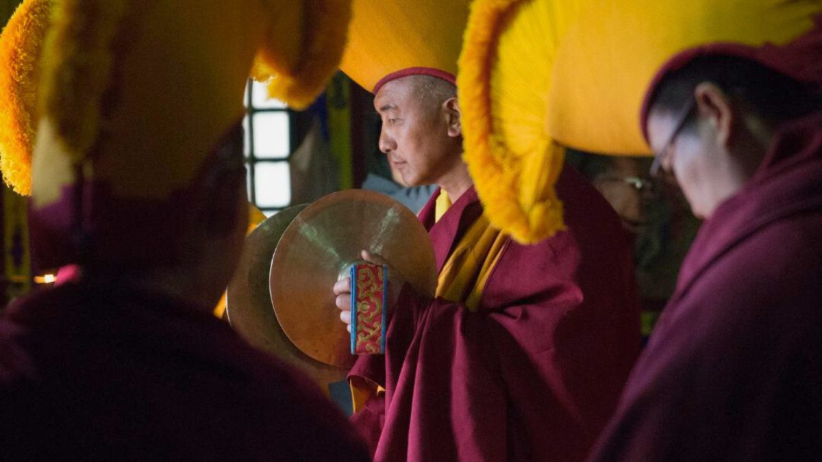 Monks in traditional robes and large yellow hats participate in a ceremony, one holding a drum while others are partially visible in the background.
