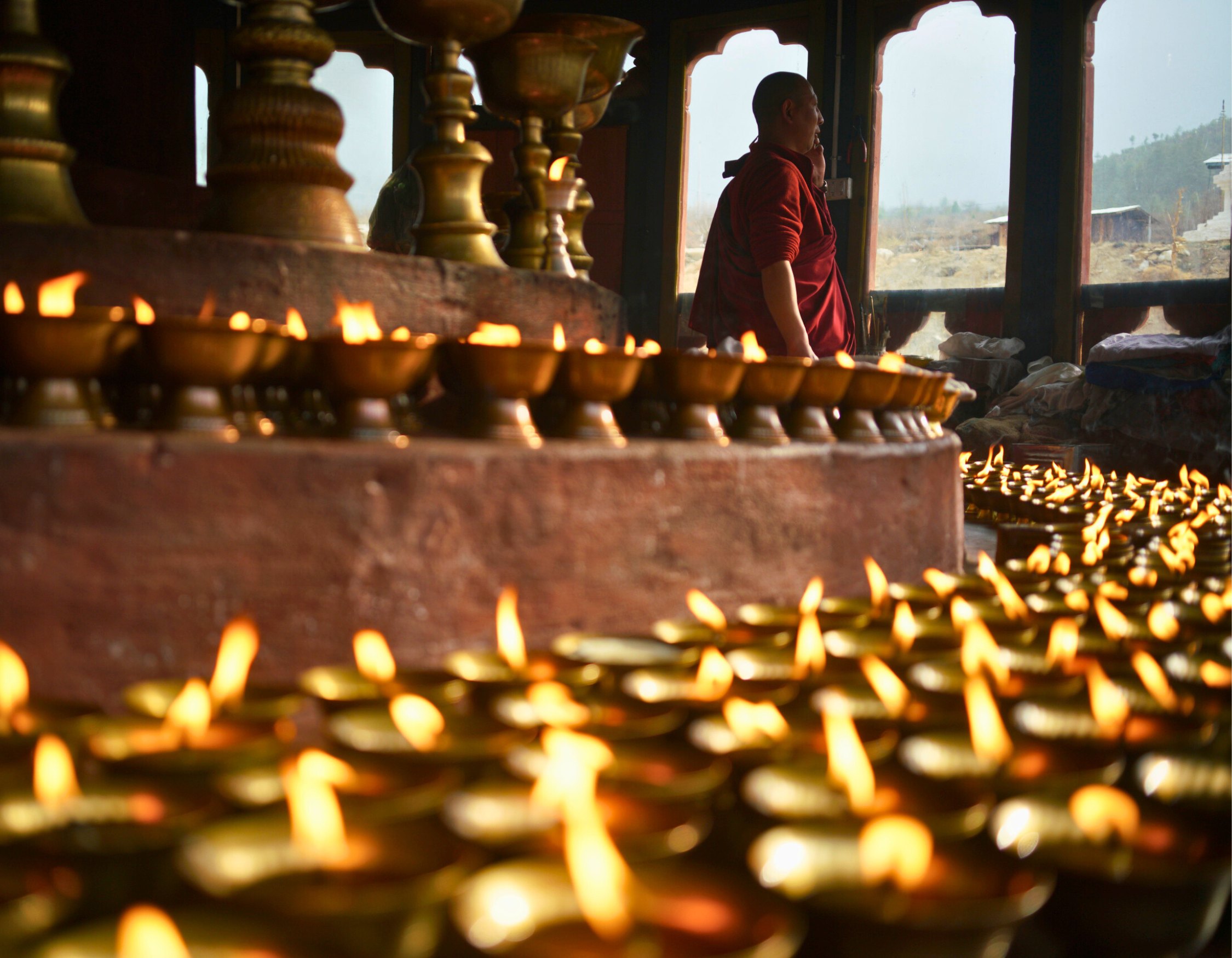 A monk stands among rows of lit oil lamps in a serene temple setting. The warm glow of the flames illuminates the surrounding area, creating a peaceful atmosphere.