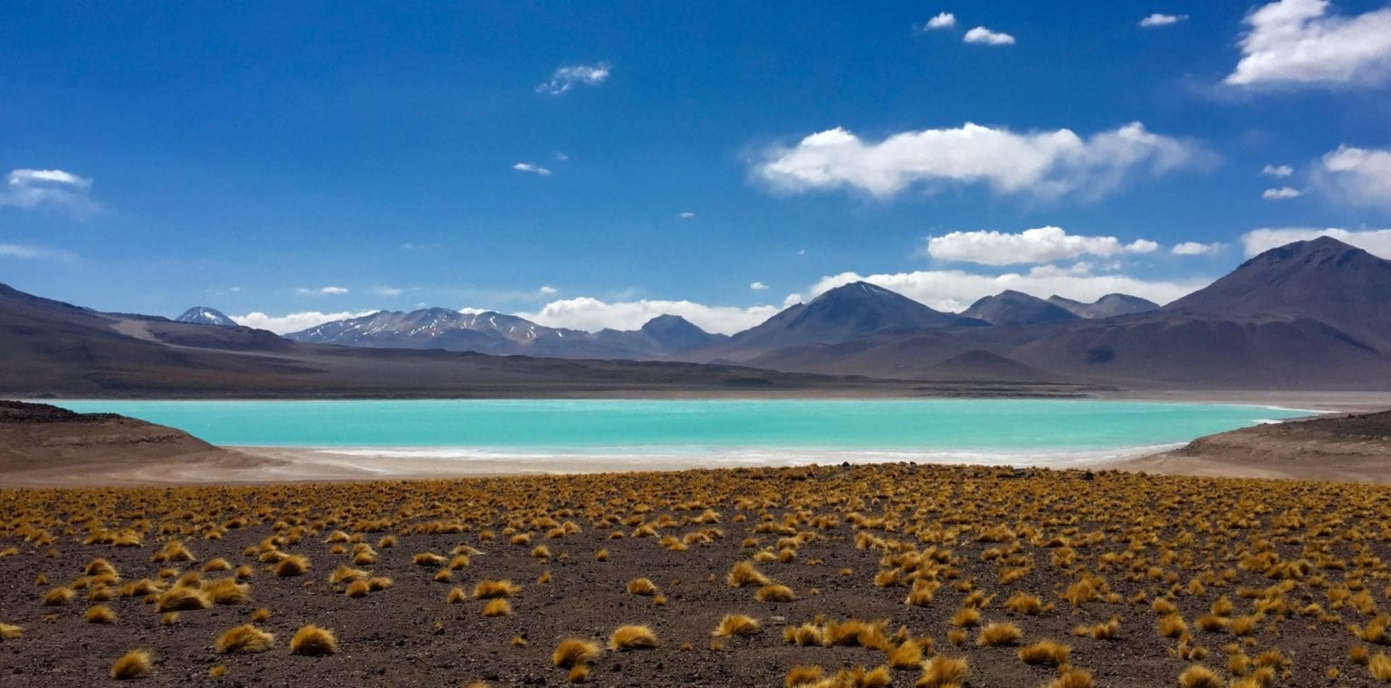 Vast landscape featuring a bright turquoise lake surrounded by arid land and sparse yellow grasses, with distant mountains under a partly cloudy blue sky.