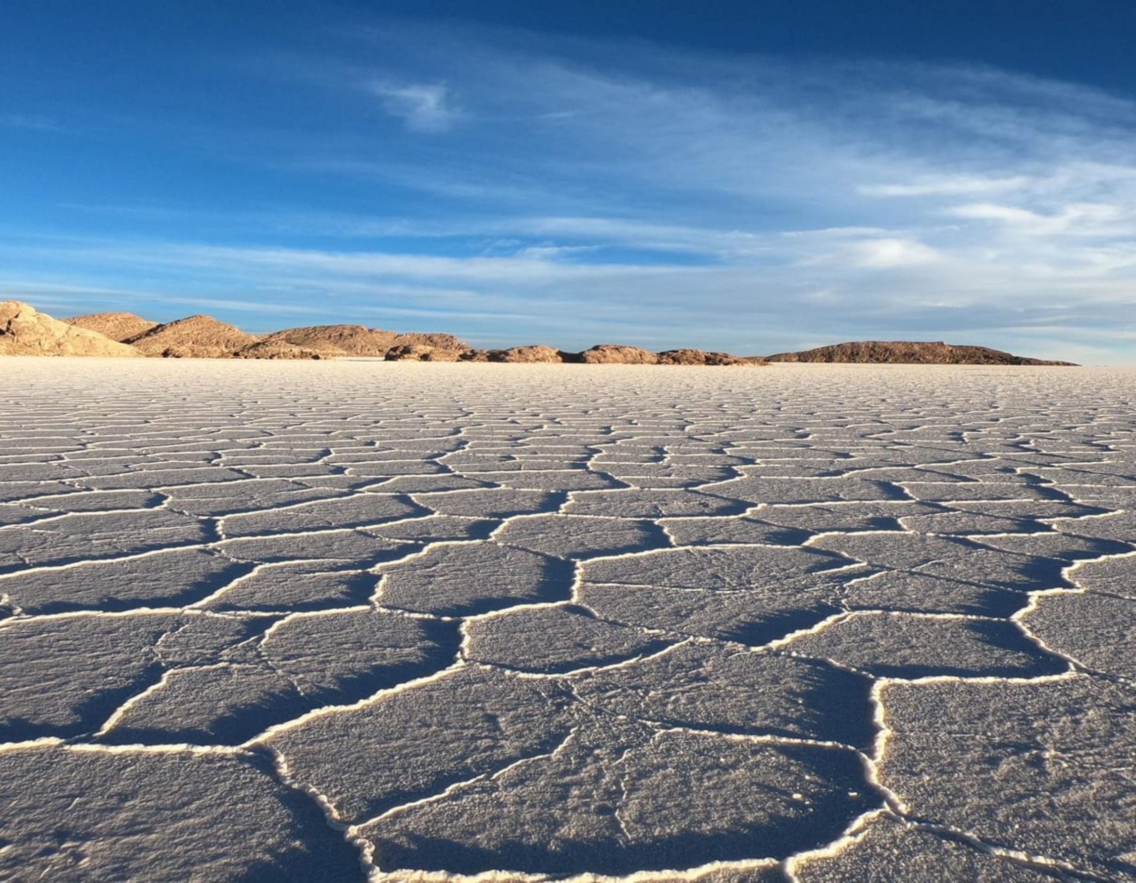A vast, flat salt flat with a cracked surface pattern stretches toward distant hills under a blue sky with wispy clouds.