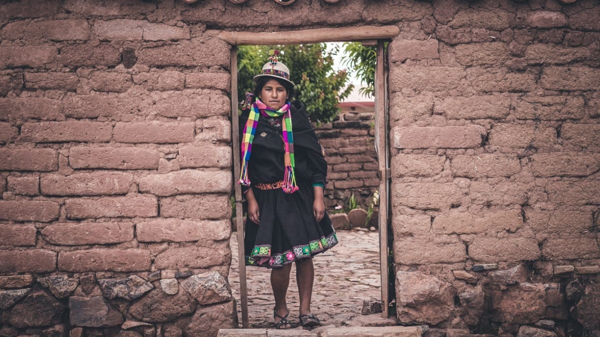 A young woman stands in the doorway of a mud-brick house, wearing traditional clothing with colorful accents and a hat. She has a serious expression and is surrounded by a rustic, rural landscape.