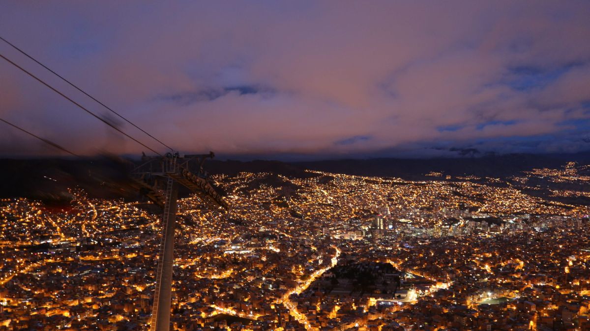 A panoramic view of a city at night, illuminated by countless lights, with a cable car line extending across the scene and dramatic clouds in the sky.
