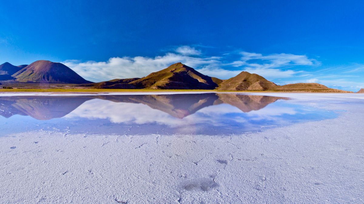 A vast salt flat reflects distant mountains under a clear blue sky, with scattered clouds floating above. The salt surface shows cracked patterns, adding texture to the serene landscape.