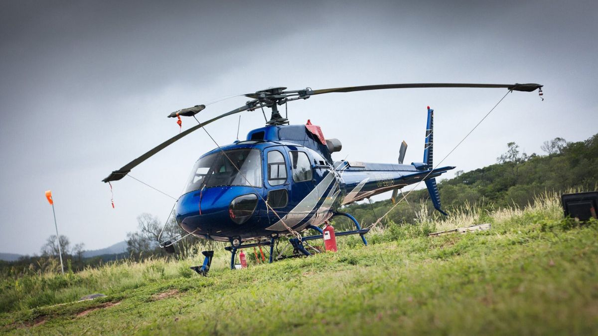 A blue and white helicopter is parked on a grassy field, with its rotors stationary. There are orange flags in the background, and a fire extinguisher is visible near the helicopter. Overcast skies create a moody atmosphere.