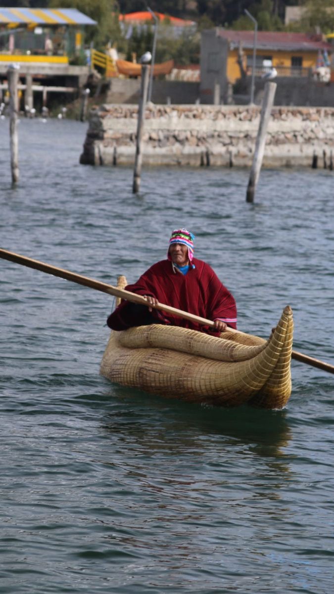 A person in traditional attire rowing a large, woven reed boat on a tranquil lake.