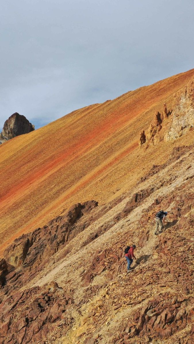 Two hikers ascend a steep, rocky mountainside characterized by vibrant orange and brown earth tones. The landscape is rugged, with a dramatic slope and cloudy skies above.
