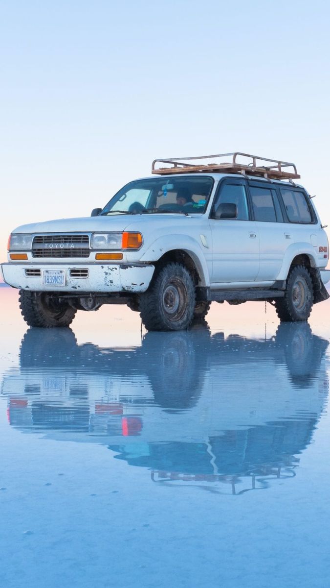 A white Toyota SUV parked on a reflective salt flat, showcasing its rugged tires and roof rack. The scene captures a clear sky above, enhancing the vehicle's sleek reflection on the wet surface.