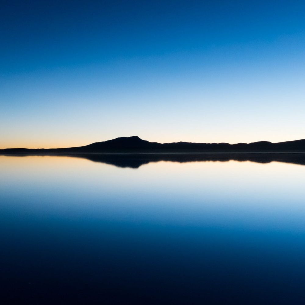 A tranquil lake reflects a gradient sky from deep blue to soft orange at the horizon, with distant mountains silhouetted along the water's edge.