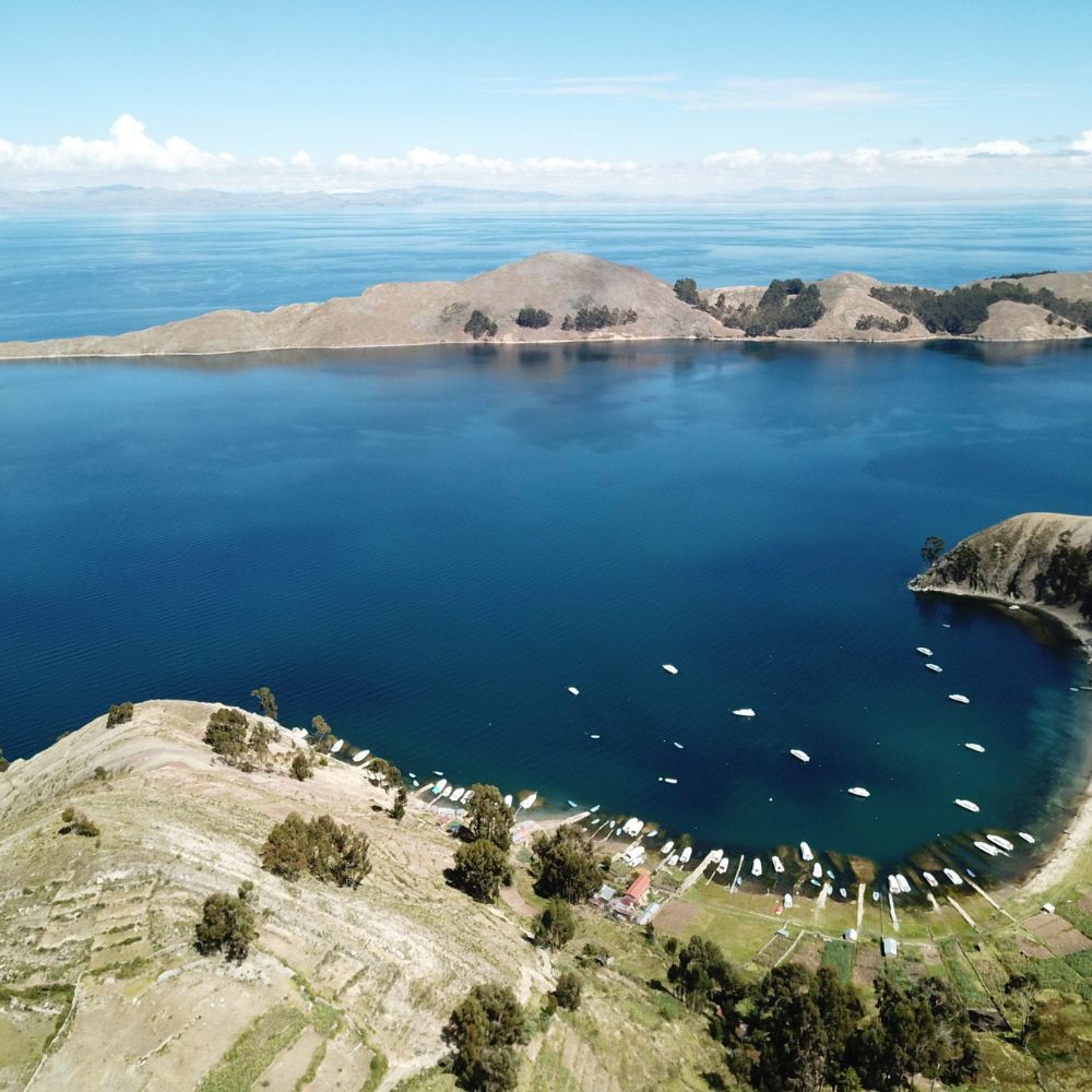 Aerial view of a serene lake surrounded by hills. Small boats are moored along the shore, with patches of greenery and farmland visible on the land. The clear blue water reflects the sky, creating a tranquil landscape.
