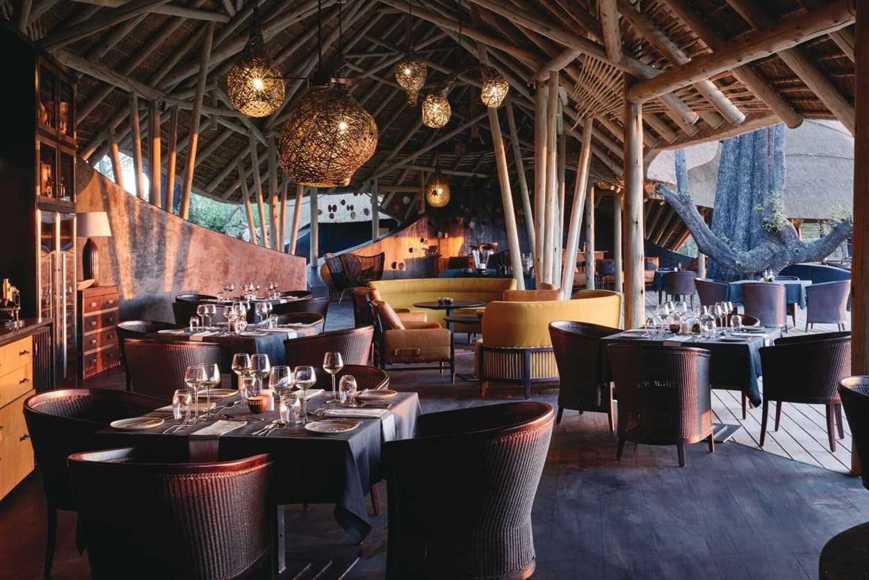 A stylish restaurant interior featuring wooden beams, a thatched roof, and modern furnishings. Elegant dining tables are set with glassware, while cozy seating areas create a warm atmosphere. Unique hanging light fixtures add to the ambiance.