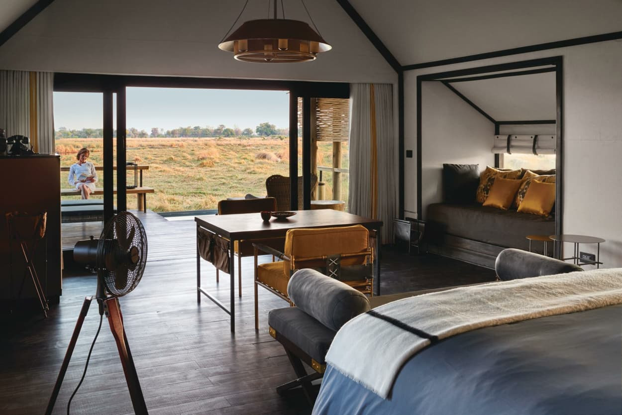A stylish interior of a bedroom is visible, featuring a large bed, a wooden dining table, and a cozy seating area. Large glass doors open to a view of a field, where a person sits outside, enjoying the landscape. The room is decorated with warm tones and modern furnishings.