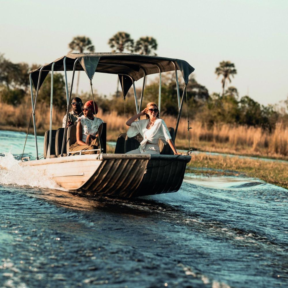 A small boat glides through calm waters, with three women onboard enjoying the ride. Lush greenery and palm trees are visible in the background under a clear sky.