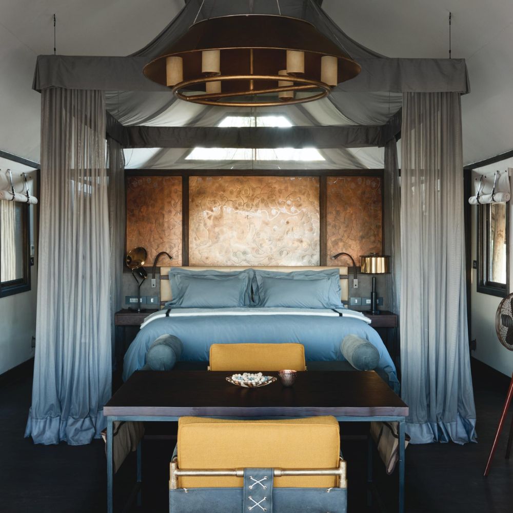 A stylish bedroom featuring a large bed with blue linens, surrounded by sheer curtains. A wooden dining table with a yellow chair sits in the foreground, and decorative elements are present on the walls. Warm lighting creates an inviting ambiance.