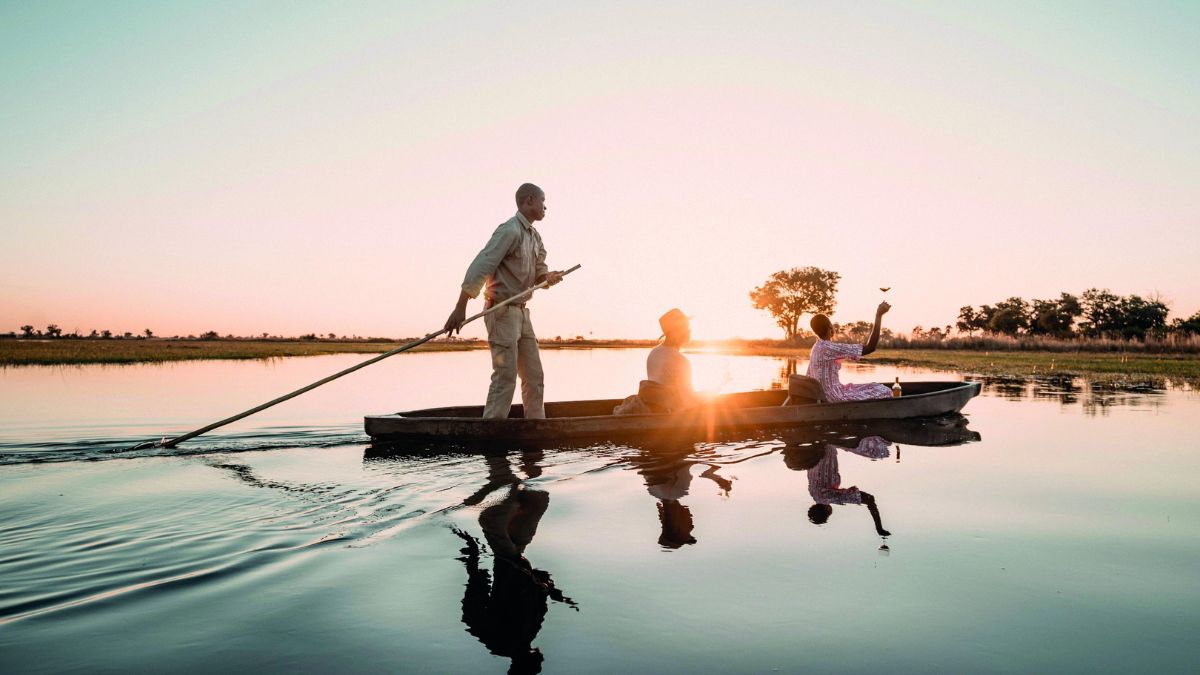 A group of three people is rowing a small boat across a tranquil river at sunset. The sun casts a warm glow, reflecting on the water's surface. One person is standing with a paddle, while another sits calmly and the third waves cheerfully. Trees line the riverbank in the background.