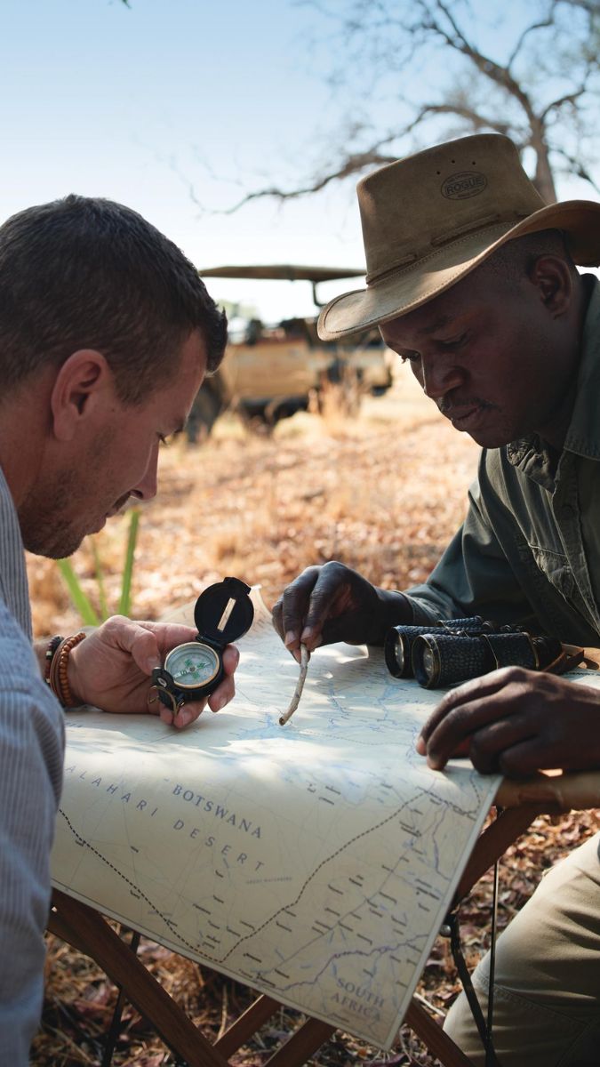Two men examine a detailed map outdoors, with one holding a compass and the other pointing at a location. They are surrounded by trees and safari vehicles in the background. One man wears a hat, while the other has a casual shirt.