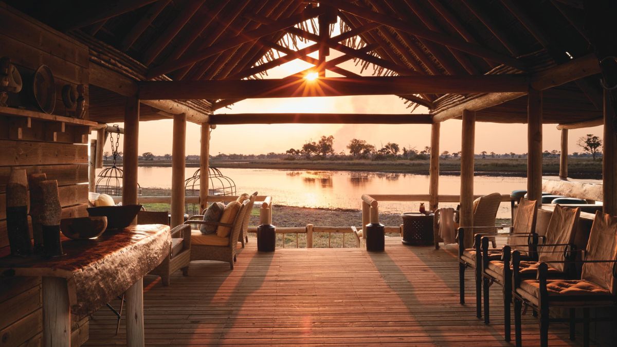 A serene lakeside patio with wooden flooring and rustic furniture, framed by a thatched roof. The sun sets in the background, casting a warm glow over the calm water and surrounding landscape.