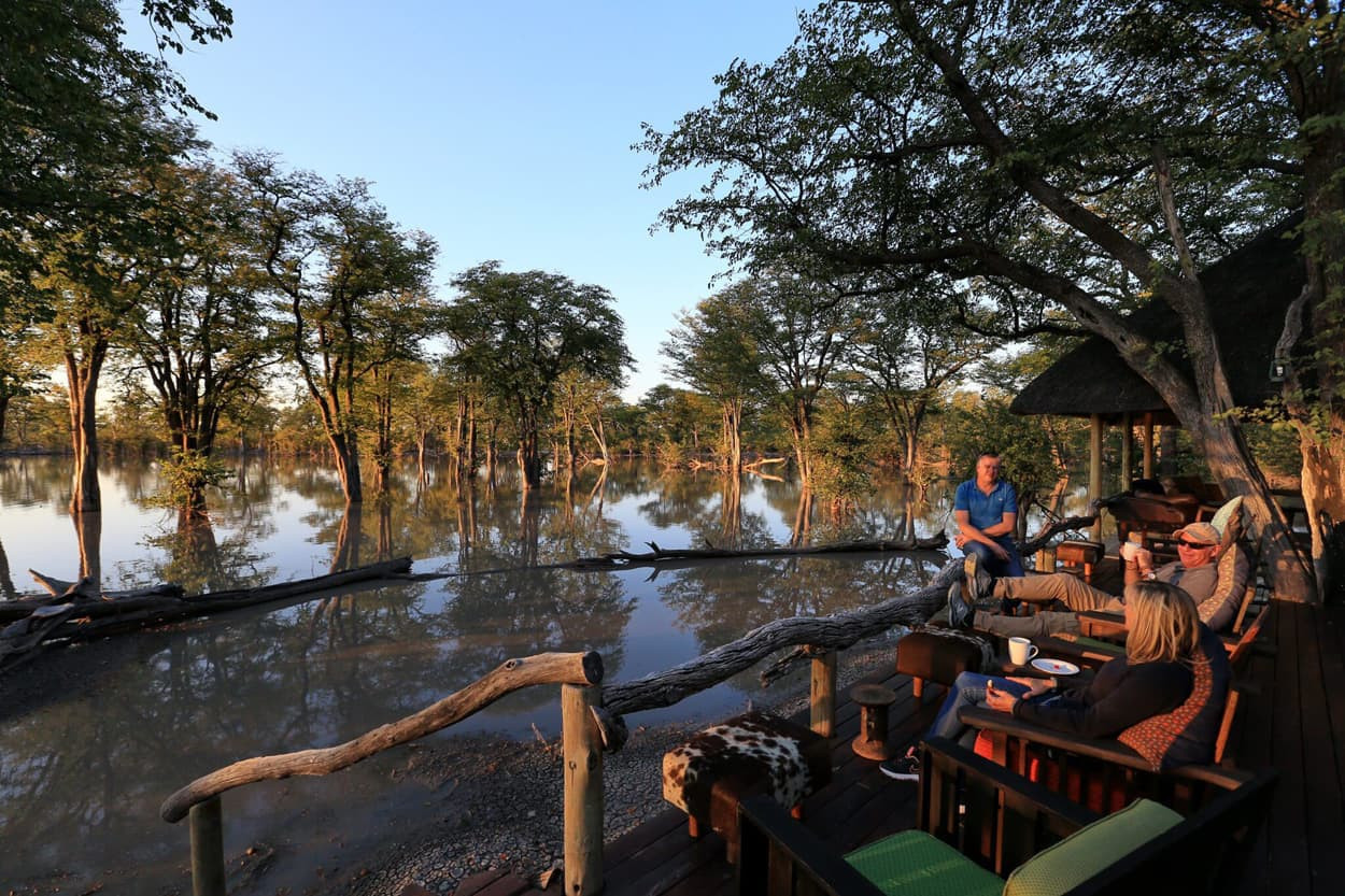 A serene lakeside scene at sunset featuring a group of people relaxing on a wooden deck surrounded by water and lush trees. The warm glow of the sun reflects off the water's surface, creating a peaceful atmosphere.