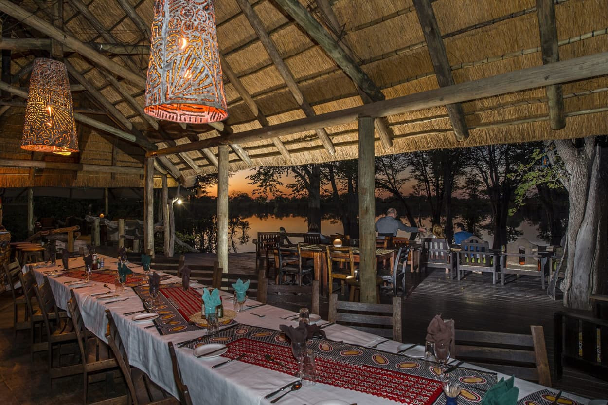 A rustic restaurant with a long table set for dining, decorated with colorful cloth and dark wooden chairs. Warm pendant lights hang from the thatched ceiling, illuminating the space. The setting overlooks a tranquil body of water and trees, with a sunset in the background.