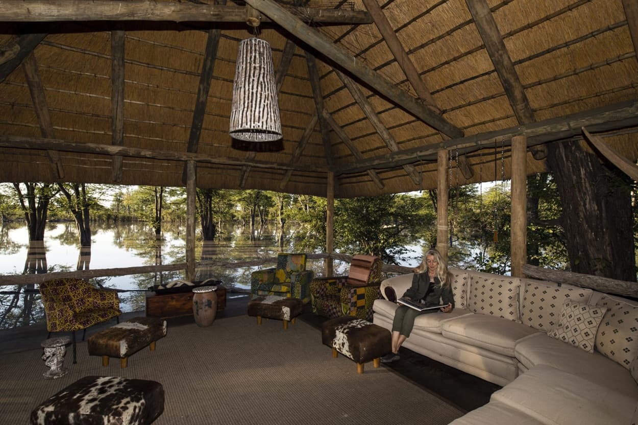 A cozy, rustic interior of a thatched-roof lodge overlooking a serene water landscape. Comfortable seating is arranged around the space, with vibrant patterned chairs and a woman seated on a sofa, reading a book. Lush greenery is visible through large windows.