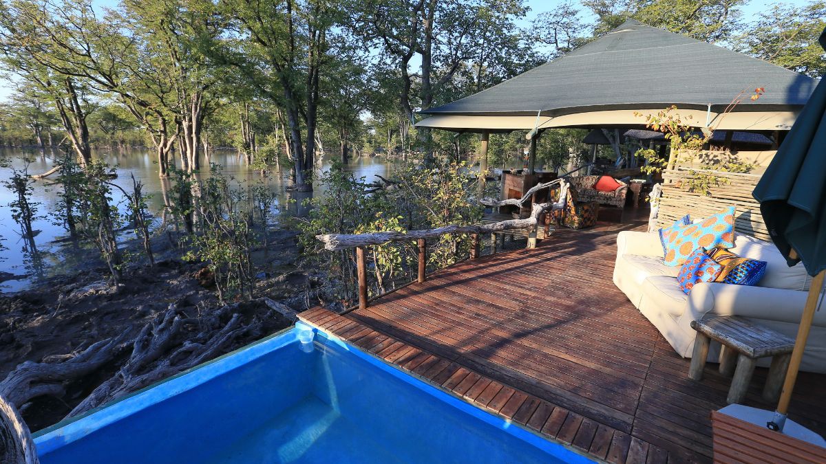 A cozy wooden deck overlooks a serene body of water, surrounded by lush greenery. A small blue pool sits on the deck, and comfortable seating with colorful pillows invites relaxation.