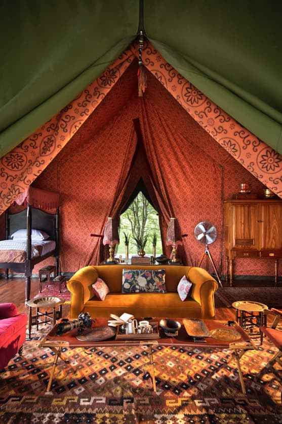 A luxurious glamping tent featuring a vibrant orange and green interior. A plush yellow sofa is centered in the space, surrounded by various decorative tables and a patterned rug. A wooden cabinet and a vintage fan are visible, along with a bed peeking from an adjoining area. Natural light filters through the entrance, illuminating the tent's cozy atmosphere.