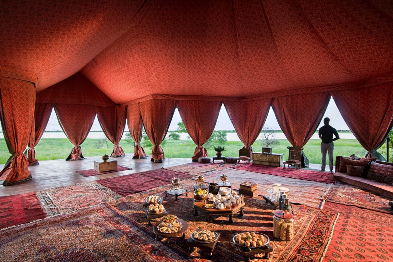 A spacious tent interior decorated with rich fabrics and intricate patterns. The room features an array of traditional dishes and drinks on low tables. Large open windows offer a view of lush greenery outside, while a person stands silhouetted by the light.