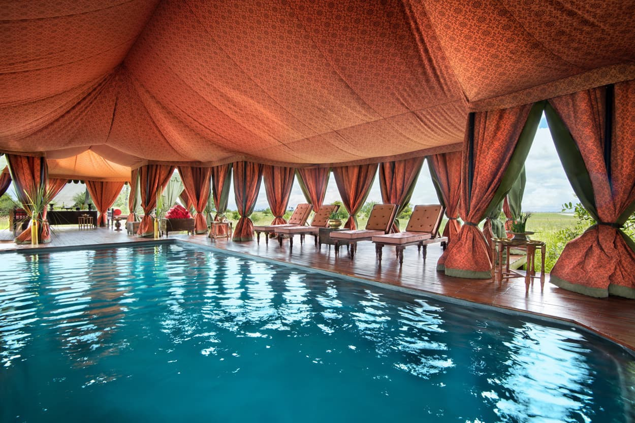 Luxurious indoor pool area with a large tent-like structure featuring vibrant orange and patterned fabric, surrounded by elegant seating and large windows offering views of greenery.