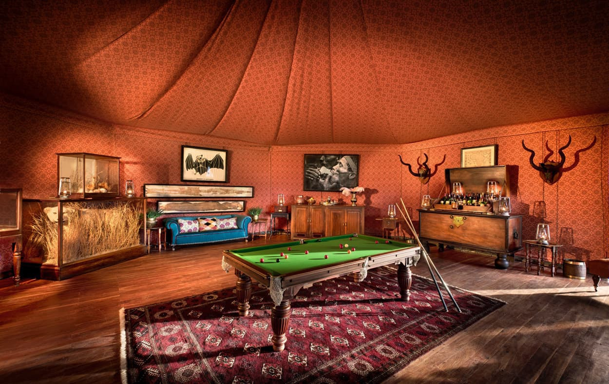 A richly decorated room with a red wallpaper featuring intricate patterns. The space includes a vintage pool table in the center, surrounded by a patterned rug. Two animal horns adorn the walls, and a wooden cabinet filled with bottles is present. Artwork and a display case with decorative items add to the ambiance.