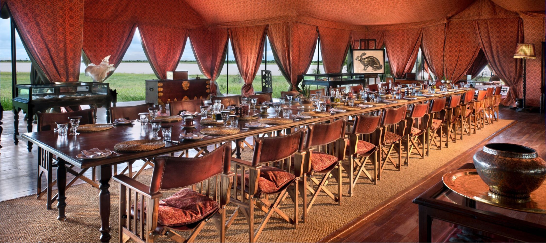 A beautifully set dining table inside a lavish tent, decorated with red and gold fabric. The long table is surrounded by wooden chairs, elegantly adorned with glasses, plates, and cutlery. Large windows reveal a scenic view of greenery outside.