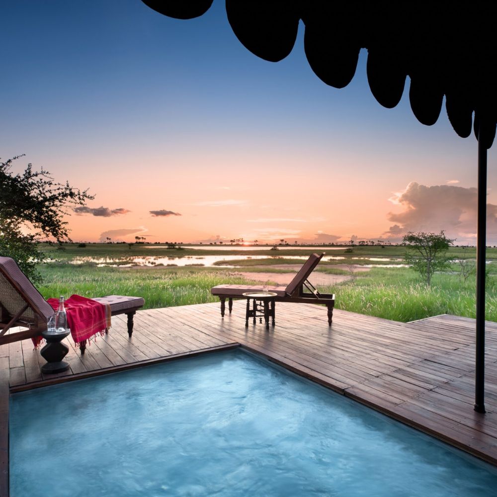 A secluded deck with a small pool overlooks a tranquil landscape at sunset. Lush green grasses are visible, with water reflecting the vibrant colors of the sky. Two lounge chairs and a red blanket add a touch of comfort to the serene setting.