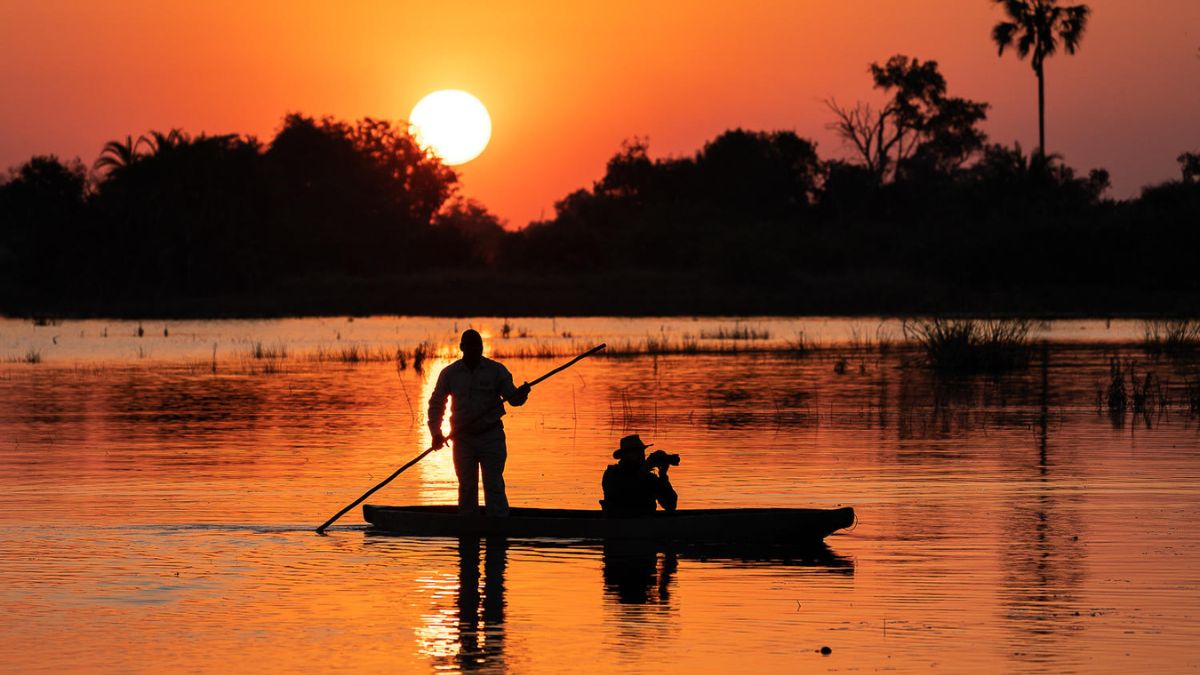 A silhouette of a person standing in a small boat with a paddle, while another person sits with a camera. The scene is set against a vibrant sunset, reflecting orange and yellow hues on the water surface, with trees and plants visible in the background.