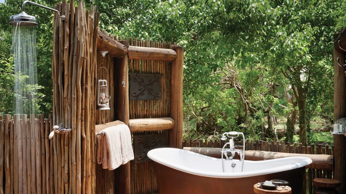 A rustic outdoor bathroom featuring a freestanding white bathtub and a shower with a natural wooden privacy fence. Lush greenery surrounds the space, enhancing the tranquil atmosphere.