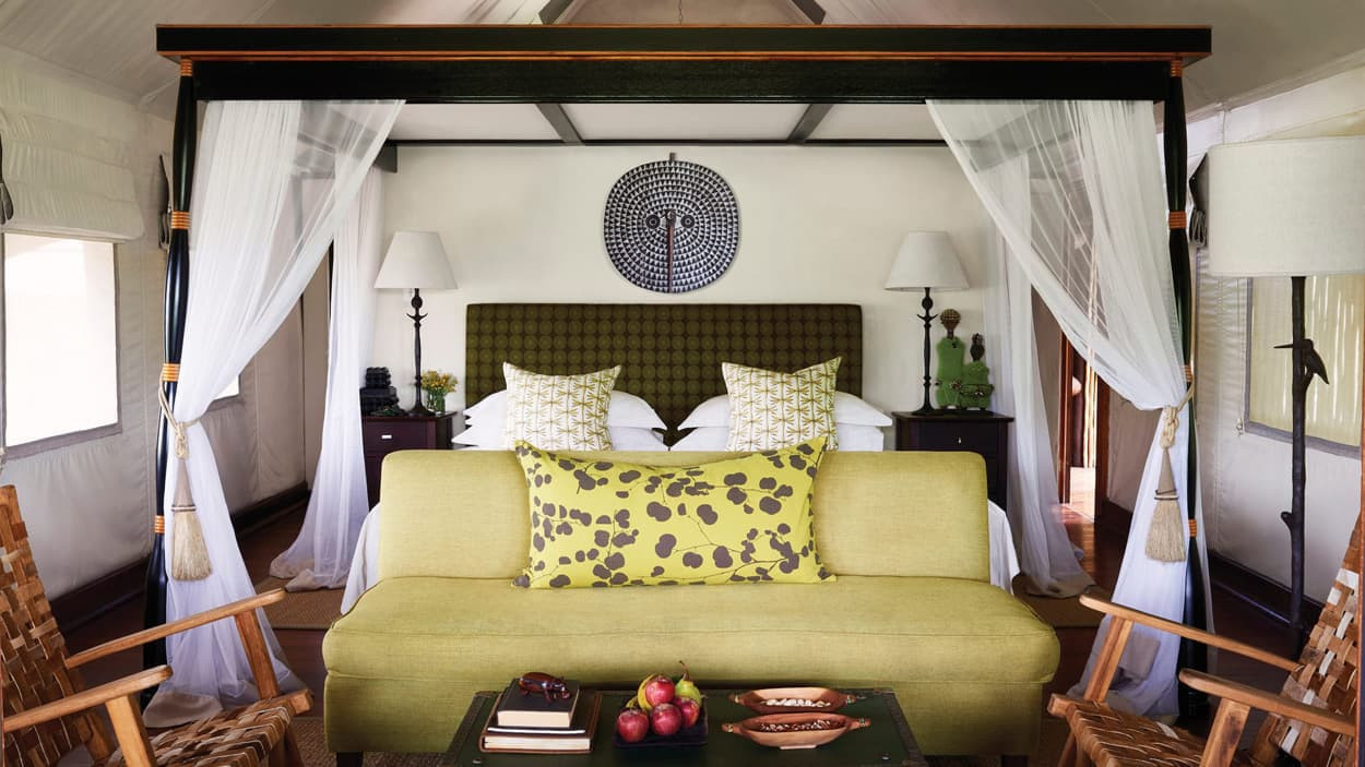 Luxurious tented bedroom featuring a four-poster bed with green and white accents, two bedside lamps, and draped sheer curtains. A bright green sofa with a decorative pillow sits in front. Decorative items and fruit are arranged on a coffee table.