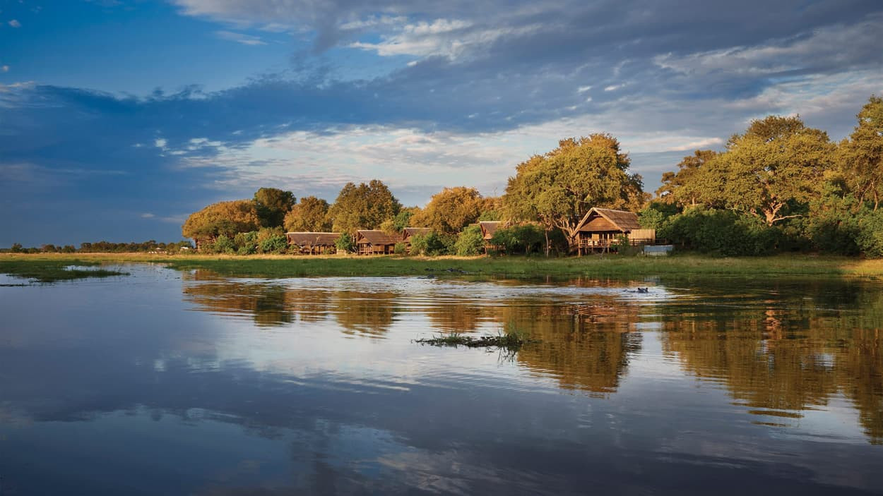 Serene river scene with wooden cabins along the shore surrounded by lush greenery and reflecting clouds in the water.
