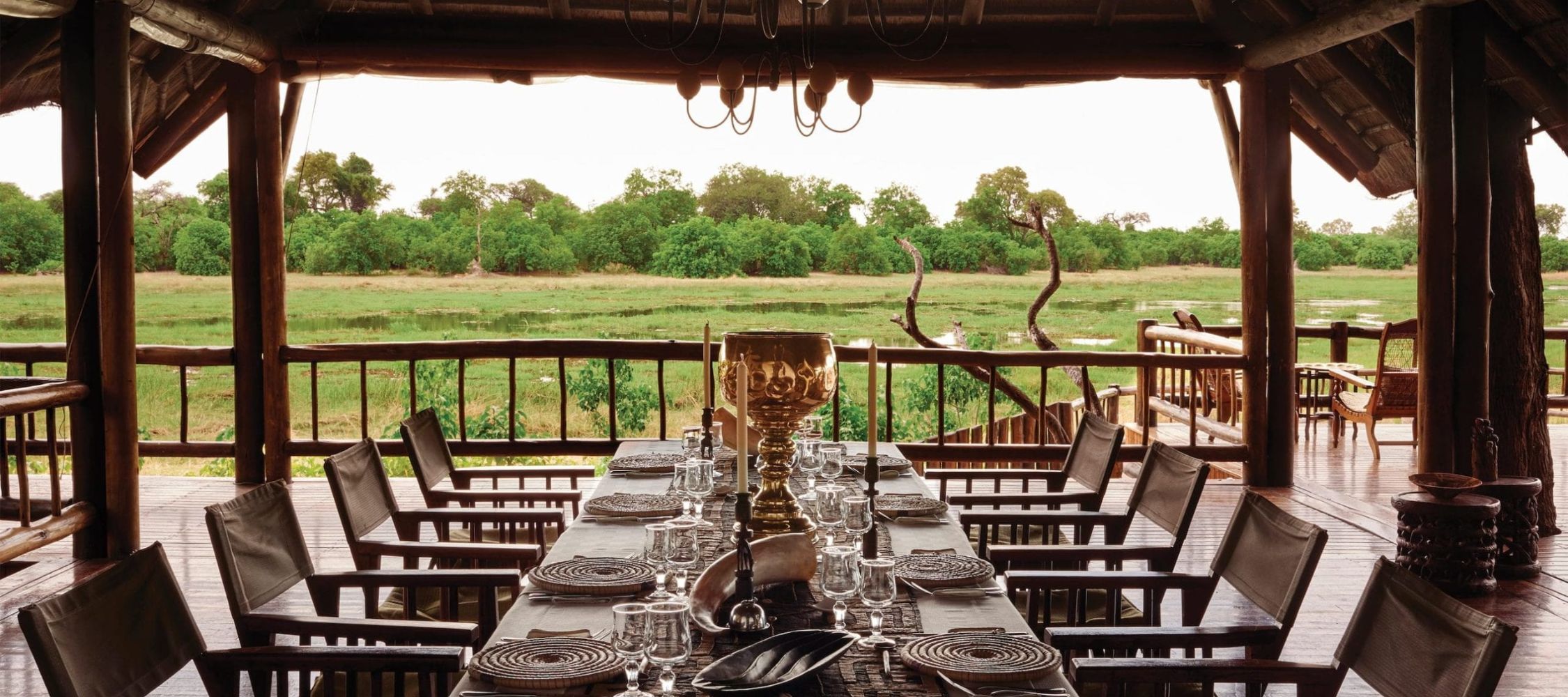 An elegant dining setup with a long table placed on a wooden deck, overlooking a lush green landscape and a river. The table is adorned with glassware, plates, and a decorative centrepiece, surrounded by comfortable chairs. Natural light illuminates the scene, enhancing the tranquil atmosphere.