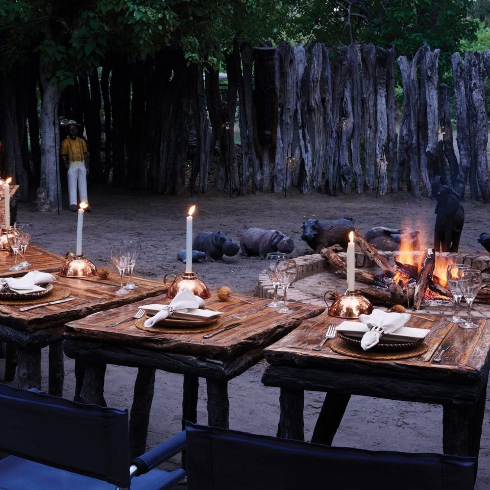 A beautifully set outdoor dining table with rustic wooden tables, adorned with elegant dinnerware and candles. In the background, a flickering fire and silhouettes of animals can be seen, framed by a rustic wooden fence.