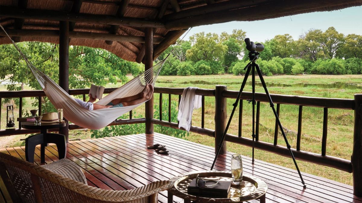 A person relaxes in a hammock on a wooden porch with a view of greenery, while a telescope stands nearby. The setting has a rustic, cozy feel with various items scattered around, including a hat and a glass.