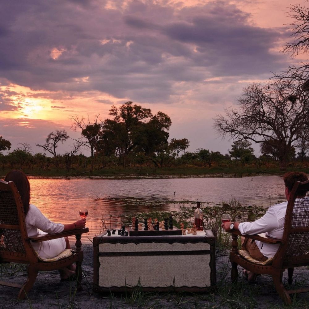Two people sit in stylish chairs by a lake at sunset, playing chess with a rose wine in hand. The landscape features trees and a tranquil water scene, creating a serene atmosphere.