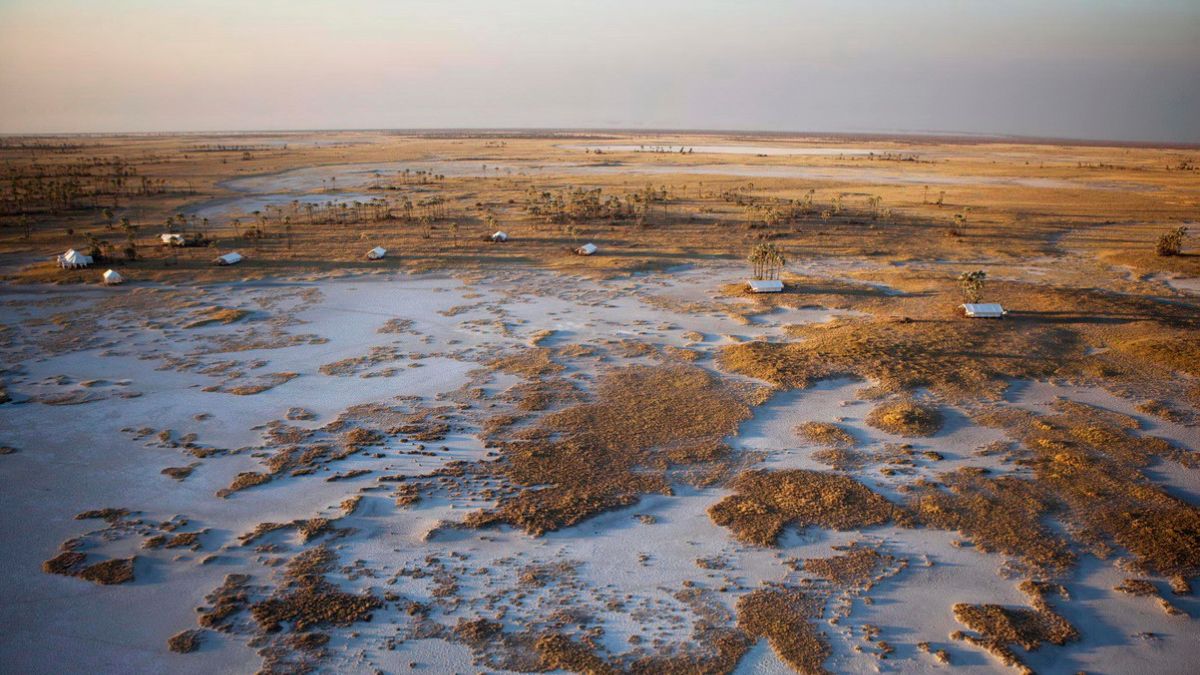Aerial view of a vast landscape featuring patches of water and dry land, with scattered small white structures and sparse vegetation under a pastel sky.