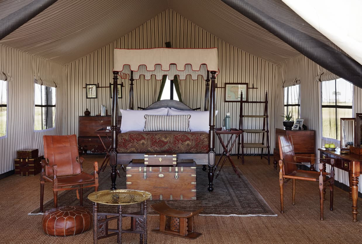 Luxurious tented interior featuring a four-poster bed with ornate bedding, wooden furniture, and a stylish rug. Natural light streams in through large windows, creating an elegant and inviting atmosphere.